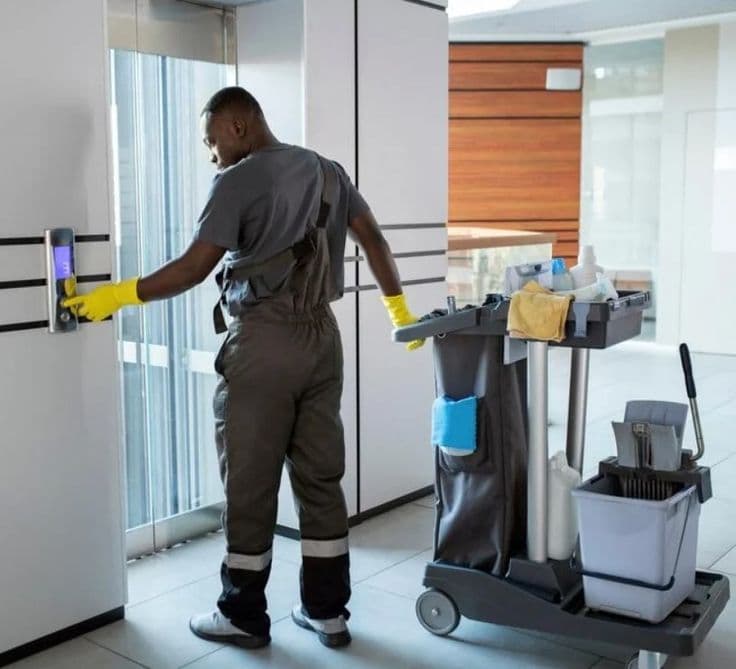 Commercial cleaner with cart beside glass elevators in a bright modern office lobby